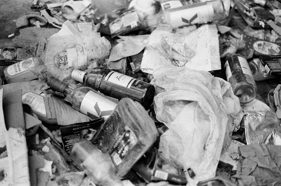 A close-up view of a densely packed collection of household waste materials scattered across a surface, consisting mainly of crumpled and torn paper, cardboard packaging, and plastic bottles. The paper items vary in texture, some appearing smooth while others are wrinkled or shredded, with a mix of plain and printed surfaces. Several transparent plastic bottles with visible labels and caps are strewn across the pile, exhibiting glossy textures and slight distortions from crumpling. Cardboard boxes and packaging materials, some with printed branding, are partially flattened and layered on top of each other, adding to the cluttered scene. The overall color tone of the image is neutral and muted, with shades of gray, black, and white, consistent with a monochrome depiction of waste. The background shows an indistinct environment that could be an outdoor or indoor space used for waste collection or disposal, with no visible machinery or cleaning tools present. This image from Waste Disposal Kennington highlights the typical clutter associated with private rubbish removal or on-site clearance tasks, emphasizing the need for proper waste handling and disposal practices in line with local regulations.