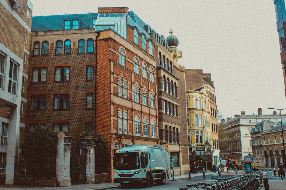 The image depicts a city street scene featuring a collection of multi-story buildings with varying architectural styles, including brick and stucco facades. Prominently positioned in the foreground is a small, light blue commercial waste collection vehicle parked alongside the curb, its doors and rear closed. To the left, a gated entrance with brick pillars and iron fencing marks a private property boundary, partially obscured by low shrubs. The street is lined with black bollards and bicycle racks, and in the background, there are other vehicles, street lamps, and additional historic and modern buildings. The overall atmosphere is daytime with natural lighting, highlighting the textures and colors of the buildings’ materials, including the reddish-brown bricks, cream-colored finishes, and the blue roofing. This scene visually supports the concept of independent rubbish collection and private waste handling services in urban environments, aligning with waste disposal solutions offered by Waste Disposal Kennington.