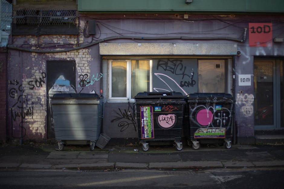 The image depicts three large waste collection bins positioned along a pavement outside a building in an urban environment. The leftmost bin is grey, made of durable plastic with a matte finish, and appears to be a standard trash or recycling container on wheels. To its right are two black bins, also on wheels, with a similar plastic construction and matte finish, each featuring colorful stickers and graffiti, including a prominent pink heart symbol and various tags. The bins are placed directly in front of a weathered building facade that has exposed brickwork and a painted surface with numerous graffiti tags and spray paint markings in different colors. Behind the bins, there is a small, white-framed window with closed blinds, and to the right, a door with glass panes also covered with graffiti. Above the scene, a narrow strip of horizontal pipes or cables runs across the wall, and the overall lighting suggests natural daylight with some shadowing, highlighting the urban, slightly neglected appearance of the setting. This scene illustrates a typical private waste handling setup, possibly related to independent rubbish collection services like Waste Disposal Kennington, catering to local businesses seeking alternative clearance options.