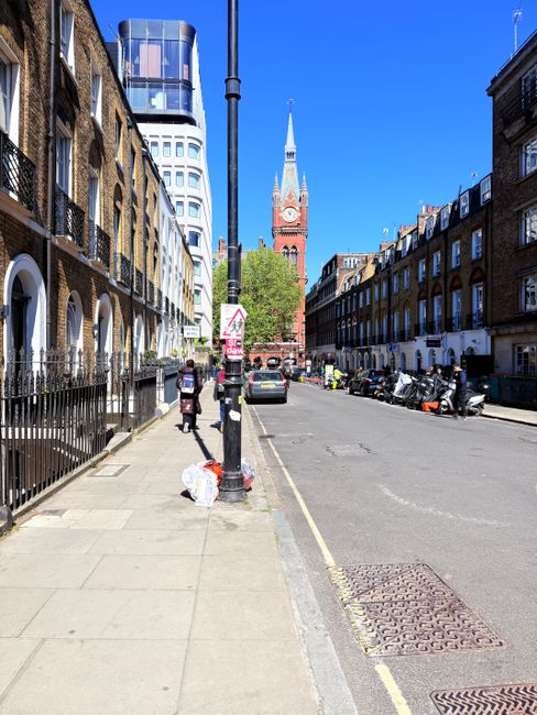A wide view of a city street in London on a clear, sunny day, with a mix of old and modern buildings lining both sides of the road. On the left side, there are brick terraced houses with arched windows, and a black metal fence separates the sidewalk from the front gardens. A person is seen walking past a parked white plastic bag on the pavement near a streetlamp, which has a pedestrian crossing sign attached. Several parked motorcycles and a few cars are lined along the right side of the street. In the background, a prominent red clock tower with a pointed spire and a large clock face rises above the surrounding buildings, with the modern glass and steel high-rise in the distance further emphasizing the urban environment. The scene appears to be in a busy, well-maintained area suitable for private waste collection or rubbish removal services, with Waste Disposal Kennington potentially involved in managing waste here, as inferred from the context of rubbish removal service relevance.