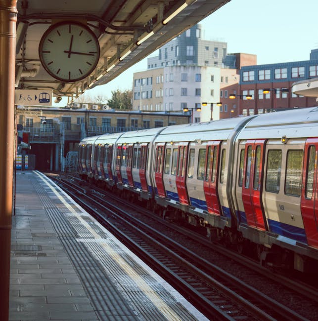 A photograph of an urban train station platform showing a modern passenger train with a sleek, metallic finish in red, blue, and silver livery parked alongside. The train has multiple carriages with large rectangular windows, and the platform's textured surface features tactile paving strips near the edge for safety. Above, an analog clock with a white face and black hands indicates the time, mounted beneath an overhanging roof supported by metal brackets. The station backdrop includes a mix of residential and commercial buildings showcasing various architectural styles, with high-rise structures visible in the distance. The scene is illuminated by natural daylight, casting soft shadows on the platform and trains, with a clear sky in the background. The image reflects a typical urban railway environment, often associated with city commuting and public transit services, and subtly supports the context of independent or alternative public transport options available in city areas.
