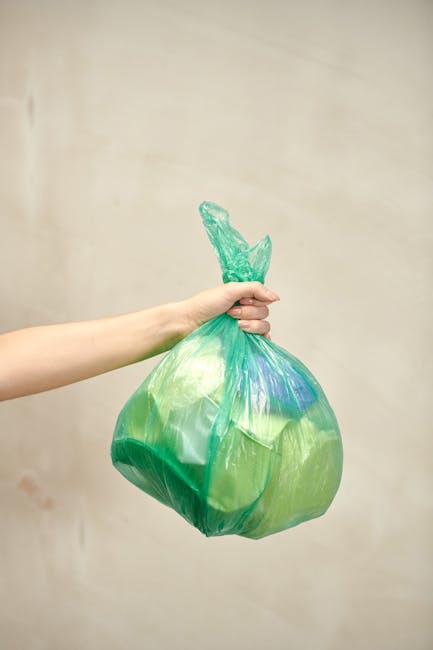 A person's hand and forearm are visible, holding a tightly knotted green plastic rubbish bag with a slightly crinkled surface and a mix of light and dark green shades, along with some blue and yellow tints. The bag appears full and is being held against a plain, neutral beige background, suggesting an indoor setting. The hand grips the bag near the top, with fingers wrapped around the knot, which is twisted and slightly frayed. The skin tone of the hand is light, with a smooth texture. The scene conveys the act of waste collection or disposal, which ties into services related to rubbish removal and alternative waste handling provided by Waste Disposal Kennington. The lighting is soft and even, emphasizing the crinkled texture of the plastic and the vibrant colour of the bag, with no shadows or reflections visible. This image exemplifies the type of waste material collected in private rubbish collection services, such as bagged household waste suitable for curbside pick-up or on-site clearance, and emphasizes the importance of proper waste containment for environmentally responsible disposal. The overall composition maintains a neutral, professional tone appropriate for informational or service-related content about rubbish management options.