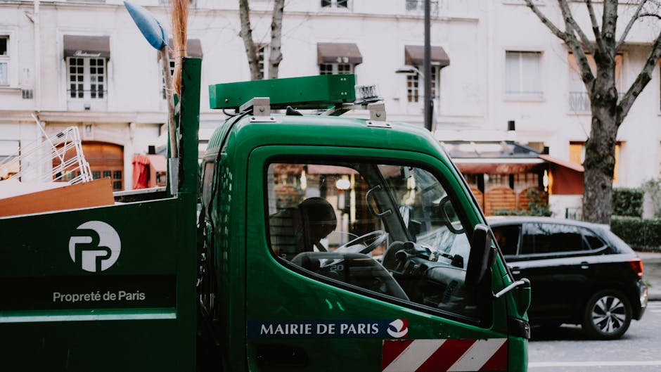 A close-up view of a green waste collection truck belonging to the Mairie de Paris, displaying the logo and the phrase 'Propreté de Paris' on its side. The truck is parked on a residential street with several leafless trees and white apartment buildings in the background. The vehicle's cab is visible, showing the steering wheel and dashboard, while the side mirror and part of the cargo area can also be seen. The environment appears to be an urban setting, possibly during daytime, with an alternative waste handling service indicated by the distinct green vehicle used for rubbish collection and disposal.