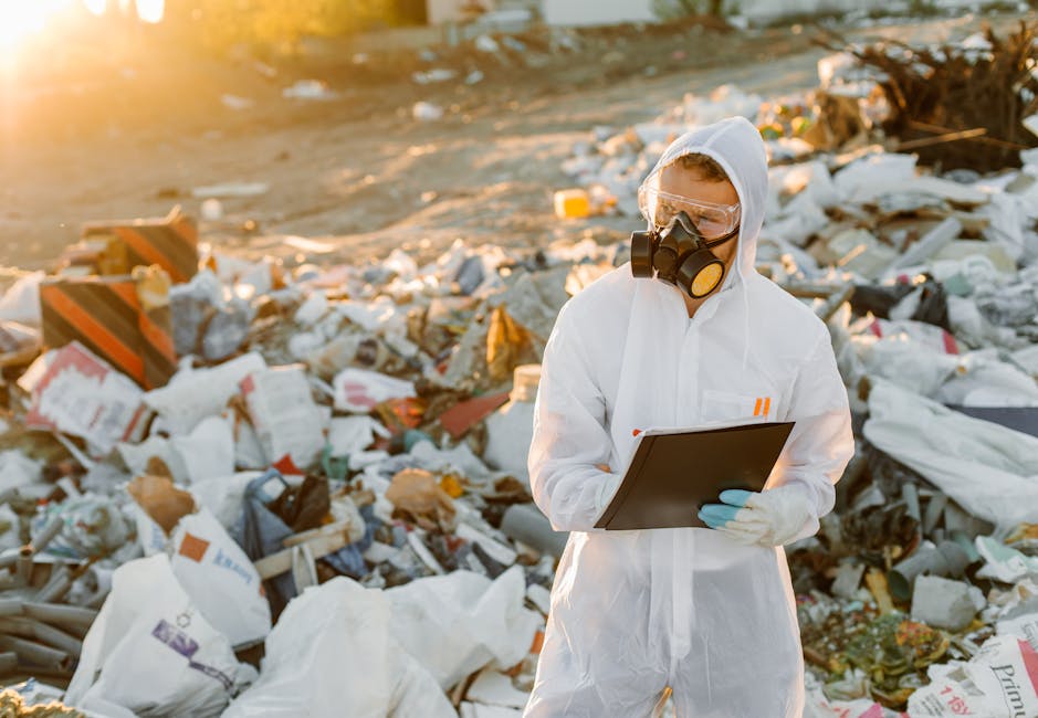 A person dressed in white protective coveralls, wearing a full-face respirator mask and blue gloves, stands amidst a large and chaotic pile of mixed waste and rubbish on an outdoor surface. The waste includes plastic bags, cardboard boxes, foam, and various discarded materials, some partially crushed or torn. The individual is holding a black clipboard and appears to be inspecting or documenting the scene, with a focused posture. In the background, the environment is illuminated by natural sunlight, casting warm tones across the scene, and the area looks like an open dump or illegal waste site. This image conveys the concept of waste management, illegal dumping, or environmental inspection, relating to services that support independent rubbish collection or on-site clearance, as provided by Waste Disposal Kennington.