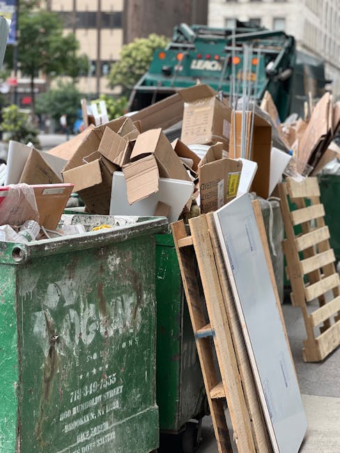 A large green waste disposal bin filled with various types of rubbish, including flattened cardboard boxes, loose cardboard pieces, and discarded packaging, situated on a city street. The cardboard is of different sizes, some corrugated, and others smooth, with visible printed labels and brown kraft surfaces. Several wooden pallets, some partially broken, lean against the bin, and a white flat-screen television is propped upright on the ground, partially obscured by the pallets. In the background, a collection vehicle with a hydraulic lifting mechanism is parked, along with urban buildings and a few trees, indicating an environment suitable for independent waste collection or on-site clearance services provided by Waste Disposal Kennington. The scene is well-lit with natural daylight, highlighting the textures of the cardboard and the metal surface of the bin, illustrating typical waste management activity in a commercial or residential street environment focused on rubbish removal and disposal.