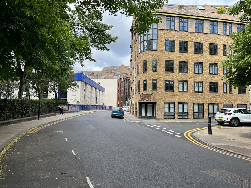 A street scene showing a curved road with double yellow lines along the curb, adjacent to a multi-storey brick building with numerous black-framed windows, some of which are open, on the right side of the image. To the left, there is a sidewalk bordered by a black metal fence, behind which lush green trees with dense foliage extend overhead, partially shading the pavement. In the background, a modern structure with blue and white panels and decorative reliefs is visible, alongside additional buildings of mixed architectural styles. Two parked cars, one dark-colored and one white, are on the right side of the street, and a person is walking along the sidewalk. The sky is overcast with grey clouds, creating a subdued natural light that illuminates the scene evenly. The environment suggests an urban area with on-site waste or rubbish removal activity, supported by the tidy appearance of the street and the presence of waste management services like Waste Disposal Kennington.