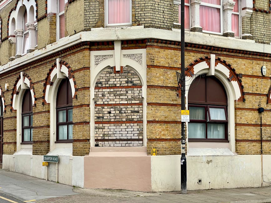 A corner of a multi-story Victorian-style brick building with a light brown and yellow brick facade, featuring arched windows with dark brown frames and white decorative trim. The ground level has three windows visible, each with curtains drawn inside. An empty near-vertical brick wall section with a decorative white arch and panel is situated between two of the windows, suggesting a possible once-fulfilled or blocked feature. A black street pole with a yellow and white sign attached is positioned in front of the building, partially obscuring the view of the rightmost window. The building sits on a concrete pavement, with a small street sign reading 'Crawford Road' at the corner, indicating the location. The scene is evenly lit with natural daylight, emphasizing the texture of the brickwork and the architectural details. This exterior context, combined with the heritage-style design, aligns with the idea of a target property for private waste removal or on-site clearance services by Waste Disposal Kennington, visible in the related webpage.
