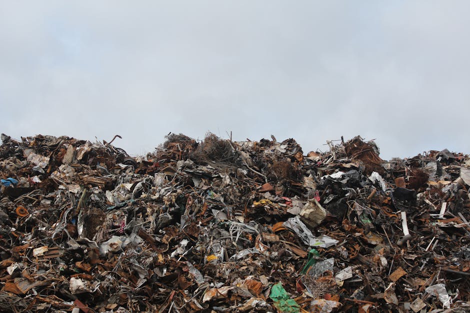 A large mound of mixed waste materials and debris dominates the lower half of the image, including twisted metal, broken plastic, discarded cardboard, and various other household and industrial refuse. The materials vary in color, with browns, greys, blacks, and sporadic splashes of brighter hues like green and blue visible among the clutter. The texture of the rubbish is rugged and uneven, with jagged edges, crumpled items, and loosely piled objects stretching across the foreground. In the background, there's a pale, overcast sky that provides a neutral, subdued backdrop to the scene, emphasizing the mess and density of the waste pile. The waste appears to be contained or piled in an outdoor environment, possibly in a designated disposal or waste collection site. The image reflects the kind of accumulated refuse that private waste disposal services, such as Waste Disposal Kennington, might handle when managing bulky or garden rubbish collections, illustrating the scale and variety of waste materials typical of alternative rubbish removal methods.