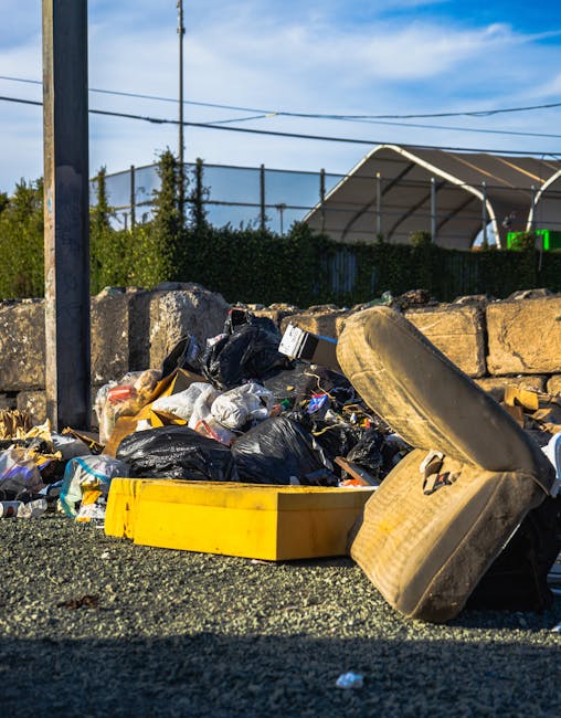 A collection of mixed household rubbish and waste materials is spilled onto the gravel surface of an outdoor area, likely near a residential or commercial property. The debris includes black plastic rubbish bags, some partially torn open, revealing assorted waste inside. There is a large, yellow plastic container positioned horizontally at the forefront, partially filled with refuse. An old, beige car seat with visible fabric upholstery and foam padding is leaning against the debris, suggesting discarded furniture as part of the waste. Behind the rubbish, a low stone wall constructed of irregularly shaped, weathered stones separates the area from the background, which includes a row of green shrubbery, a tall utility pole with electrical wires overhead, and a partially covered outdoor structure with a curved, metallic roof. The scene is illuminated by natural daylight, indicating fair weather, and the overall setting appears to be an untidy spot where private waste disposal or alternative rubbish collection services, such as those provided by Waste Disposal Kennington, might be involved.