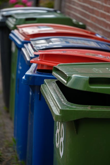 A large mound of mixed waste materials and debris dominates the lower half of the image, including twisted metal, broken plastic, discarded cardboard, and various other household and industrial refuse. The materials vary in color, with browns, greys, blacks, and sporadic splashes of brighter hues like green and blue visible among the clutter. The texture of the rubbish is rugged and uneven, with jagged edges, crumpled items, and loosely piled objects stretching across the foreground. In the background, there's a pale, overcast sky that provides a neutral, subdued backdrop to the scene, emphasizing the mess and density of the waste pile. The waste appears to be contained or piled in an outdoor environment, possibly in a designated disposal or waste collection site. The image reflects the kind of accumulated refuse that private waste disposal services, such as Waste Disposal Kennington, might handle when managing bulky or garden rubbish collections, illustrating the scale and variety of waste materials typical of alternative rubbish removal methods.