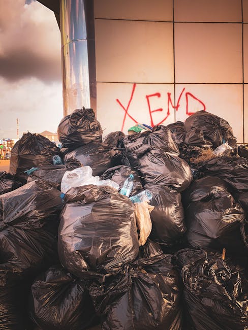 A large pile of black plastic rubbish bags, filled and tied, stacked against a modern tiled building wall. Some bags are slightly torn or bulging, revealing various waste items inside. A few discarded plastic water bottles and pieces of paper are visible on or around the bags, adding to the cluttered appearance. Behind the pile, the building features a smooth, reflective surface with square tiles in neutral tones, and there is red graffiti forming the word 'XEND' spray-painted on the wall. The scene is outdoors, with an overcast sky visible in the background, and the lighting highlights the texture of the plastic bags and surrounding environment. This image depicts an instance of private waste accumulation, potentially requiring professional rubbish removal services such as those provided by Waste Disposal Kennington, to manage non-sorted, on-site rubbish.