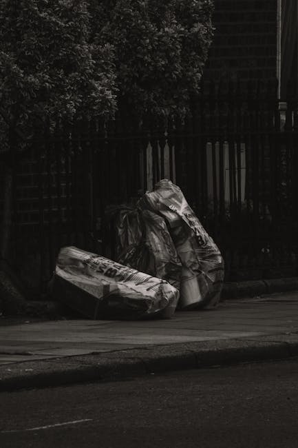 Two large black plastic rubbish bags, crumpled and partially torn, are placed on the sidewalk in front of a wooden fence with vertical slats. The bags appear to contain waste material and are positioned close together, leaning slightly against each other. Behind the fence, there are dense leafy trees or bushes, creating a dark, shaded background. The scene is captured under natural light, highlighting the textures of the plastic bags and the rough surface of the pavement. The setting suggests a typical urban outdoor environment, possibly indicating a situation of on-site waste disposal managed by independent waste removal services such as Waste Disposal Kennington, with the bags ready for collection or removal from the premises, supporting the context of alternative rubbish handling outside of municipal services.