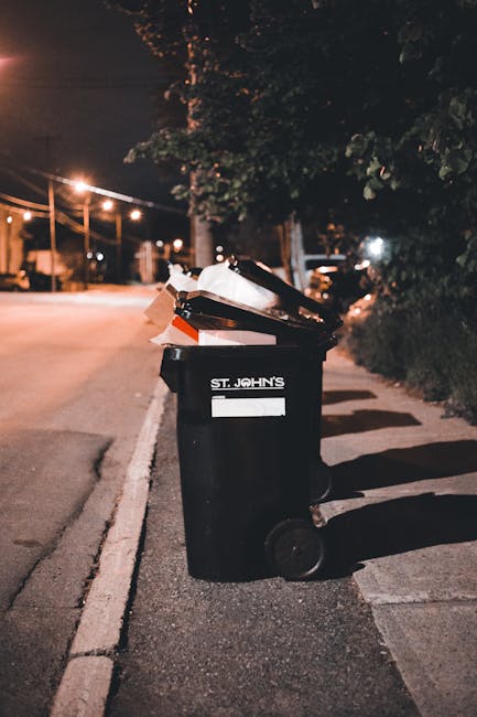 A black plastic rubbish bin with a white label and the words 'St. John's' printed on it is positioned on a sidewalk beside a curb. The bin's lid is open, revealing piles of mixed waste, including cardboard, paper, and plastic bags. The bin appears to be used for general waste collection and is situated in an outdoor urban environment during nighttime. In the background, there are streetlights illuminating a quiet street with some parked vehicles and a row of trees casting shadows, with a dark sky overhead. The surrounding area includes a narrow pavement and a hedge along the sidewalk, suggesting a residential or mixed-use area. The scene reflects a typical setting where private waste collection or rubbish removal services, such as those provided by Waste Disposal Kennington, might manage waste outside individual properties as an alternative to on-street council rubbish collection, emphasizing an independent waste handling approach.